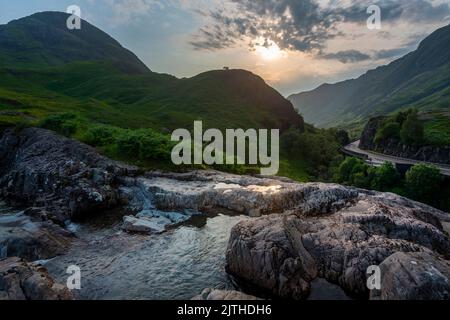 Die untergehende Sonne, über dem dramatischen Tal von Glen Coe, im Sommer, wo sich drei kleine Flüsse treffen, am Fuße der Three Sisters of Glen Coe, mit AW Stockfoto