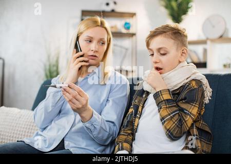 Mutter hält Thermometer und Handy, ruft den Arzt an, während sie die Temperatur ihres kranken Teenagersohnes überprüft, der mit hohem Fieber hustet und auf dem Sofa sitzt. Stockfoto