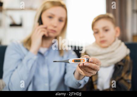 Mutter zeigt dem Kamerathermometer, nachdem sie die Temperatur gemessen hatte, während sie mit dem Handy telefoniert und einen Arzt anrief. Mutter überprüft die Temperatur ihres kranken Teenagersohnes mit hohem Fieber, der auf dem Sofa sitzt. Stockfoto