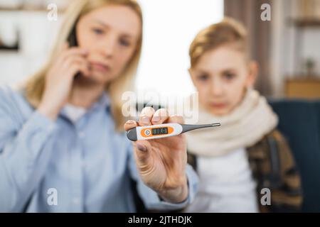 Mutter zeigt dem Kamerathermometer, nachdem sie die Temperatur gemessen hatte, während sie mit dem Handy telefoniert und einen Arzt anrief. Mutter überprüft die Temperatur ihres kranken Teenagersohnes mit hohem Fieber, der auf dem Sofa sitzt. Stockfoto