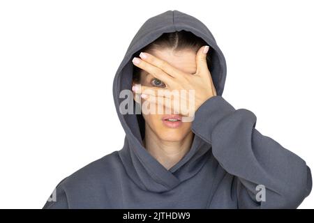 Ein Mädchen in einem Trainingsanzug schließt ein Auge mit ihrer Hand auf weißem Hintergrund, schau durch ihre Hand, ich sehe Stockfoto