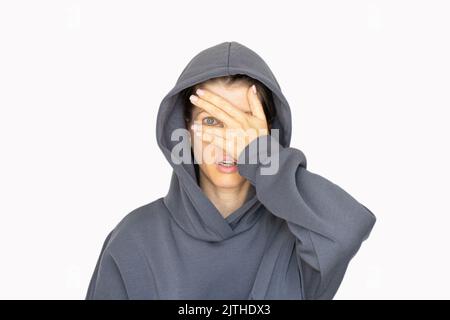 Ein Mädchen in einem Trainingsanzug schließt ein Auge mit ihrer Hand auf weißem Hintergrund, schau durch ihre Hand, ich sehe Stockfoto