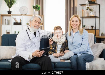 Unterstützender, aufmerksamer Arzt besucht kranken jungen Patienten zu Hause, sitzt mit Mutter, hört seinen Beschwerden zu und schaut in die Kamera. Arzt Beratung Vorschulkind zu Hause. Stockfoto