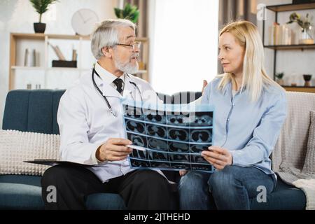 Porträt eines attraktiven bärtigen männlichen Arztes, der während des Besuches zu Hause über die Ergebnisse des Röntgenbildes mit einer Patientin mittleren Alters spricht. Stockfoto