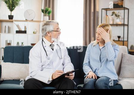 Graues Haar bärtig reifen männlichen Arzt besucht seinen hübschen mittleren Alter Patienten zu Hause. Der Therapeut notiert sich, während sich die junge Frau über schreckliche Kopfschmerzen beschwert. Stockfoto