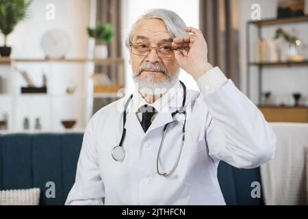 Ein nachdenklicher, gealterter Arzt in Laborkittel und Brillen blickte auf die Kamera, während er im Krankenhausbüro saß. Porträt eines qualifizierten Arztes. Stockfoto