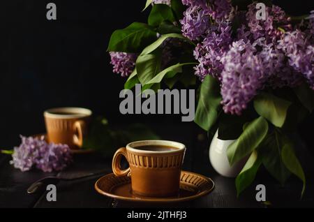 Tee am Abend mit einem Freund in gemütlicher Umgebung, braune Tassen in Unschärfe. Dunkle Taste. Teepaar mit Blumenstrauß von Fliedern auf schwarzem Holztisch. Weicher Fokus auf Fliederfluss Stockfoto