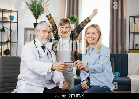 Angenehme reife männliche Arzt, im Gespräch mit konzentrierten ziemlich kaukasischen Frau über die Einnahme von Medikamenten während des Hausbesuchs des kranken Jungen, stehend mit erhobenen Händen jubeln in Gesundheit und Erholung. Stockfoto