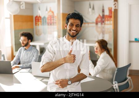 Porträt eines lächelnden indischen Geschäftsmannes, der in einem modernen Büro steht und den Daumen nach oben zeigt Stockfoto