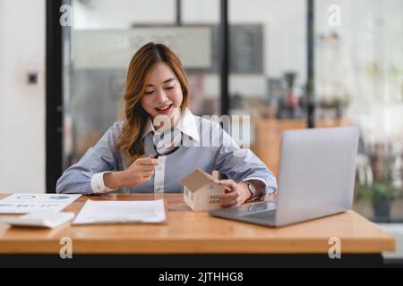 Auf der Suche nach Immobilienagentur, Immobilienversicherung, Hypothekendarlehen oder neues Haus. Frau mit Lupe über einem Holzhaus in ihrem Büro Stockfoto