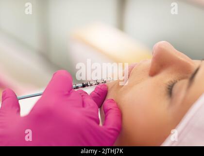 Nahaufnahme einer jungen Frau, die eine Schönheitsspritze in die Lippen bekommt. Kosmetologe machen Verfahren in rosa Handschuhe im Salon. Stockfoto