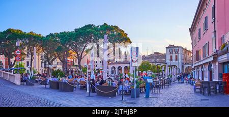 DESENZANO DEL GARDA, ITALIEN - 10. APRIL 2022: Das Abendpanorama des Alten Hafens mit Restaurants im Freien und hohen Kiefern, am 10. April in Desenzano de Stockfoto