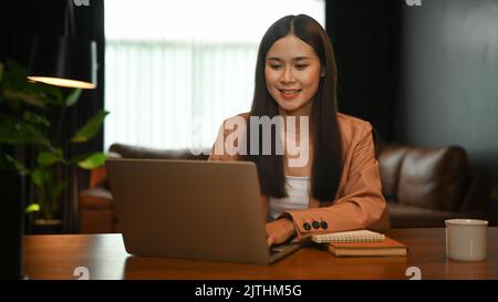 Charmante asiatische Frau, die sich das Online-Webinar auf ihrem Laptop ansieht, während sie von zu Hause aus fernarbeitet Stockfoto