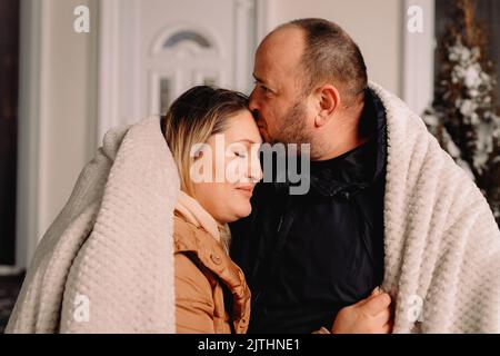 Nahaufnahme eines liebevollen Paares draußen in warmen Kleidern und einer Decke, die sich zusammenzieht und der Mann seine Frau auf die Stirn küsst. Stockfoto