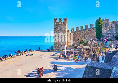 SIRMIONE, ITALIEN - 10. APRIL 2022: Die mittelalterlichen Türme und Mauern der Burg Scaligero mit einem überfüllten öffentlichen Strand am Gardasee im Vordergrund, auf AP Stockfoto