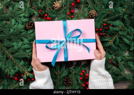 Frau Hände halten weihnachtsgeschenk oder Geschenkbox dekoriert Tannenbaum. Draufsicht auf die Hände einer jungen Frau, die ein Weihnachtsgeschenk in pi verpackt hält Stockfoto