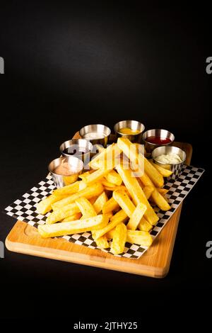 Pommes frites auf einem Holztisch, begleitet von verschiedenen Saucen auf schwarzem Hintergrund Stockfoto