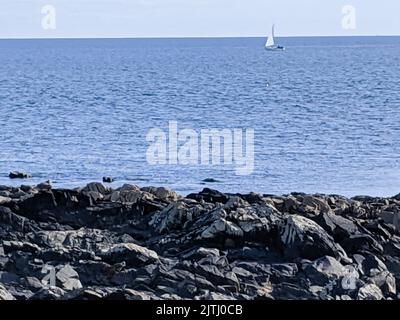 Kleine Yacht weit draußen auf dem Meer mit Küstenfelsen im Vordergrund. Stockfoto