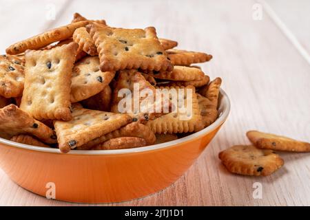 Saltine Cracker in einer orangefarbenen Schale auf einem weißen Holztisch. Tiefer Teller voller salziger Cracker mit schwarzen und weißen Sesamsamen aus der Nähe. Bereit zum Essen. Stockfoto