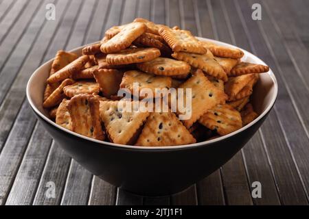 Haufen Saltine Cracker in einer Schüssel über dunklem Holzbrett Hintergrund. Schwarzer tiefer Teller voller salziger Cracker mit schwarzen und weißen Sesamkörnern. Stockfoto