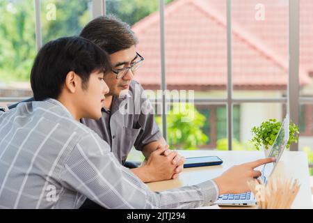 Asiatischer junger Mann lehrt älteren Vater, wie man einen modernen Laptop-Computer zusammen im Wohnzimmer zu Hause, Sohn hilft Papa, den Laptop zu verwenden für Stockfoto