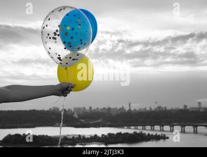 Gelbe und blaue Heliumballons in der Hand gegen den Himmel und den Fluss Dnjepr in Kiew. Farbakzent der Flagge der Ukraine. Hoffnungen auf Glück während des Kampfes auf Stockfoto