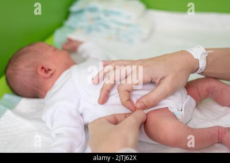 Die Hände einer Mutter, die gerade in der Entbindungsklinik geboren wurde, legen ihrem neugeborenen Baby einen Body an, nachdem sie gebadet und seine Windel gewechselt hat. Ne Stockfoto