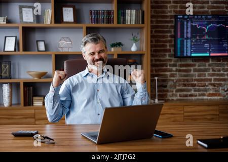 Der glückliche Geschäftsmann war von den guten Nachrichten überrascht und machte eine Ja-Geste, während er auf den Laptop im Büro schaute. Trader genießen Erfolg, feiern Stockfoto