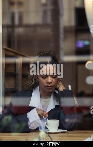 afroamerikanische Frau im trendigen Blazer, die Kaffee in der Tasse rührt, während sie hinter dem Glas im Café sitzt Stockfoto