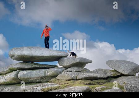 Ein Wanderer mit einer leuchtend roten Jacke, der auf einem riesigen Granitfelsen-Stapel auf dem Gipfel des Stowes-Hügels auf dem schroffen Bodmin Moor in Cornwall in den USA steht Stockfoto