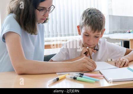 Ein Junge und ein Lehrer machen Hausaufgaben, schreiben Text in ein Notizbuch am Tisch. Eine Mutter hilft ihrem Sohn, in ein Notizbuch zu schreiben. Der Babysitter hilft beim compl Stockfoto