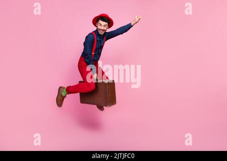 Ganzkörper-Größe Foto von jungen schönen lustigen Mann tragen roten Hut positive gute Laune Faust bis Superheld fliegen seine magische Tasche neue Abenteuer auf rosa isoliert Stockfoto