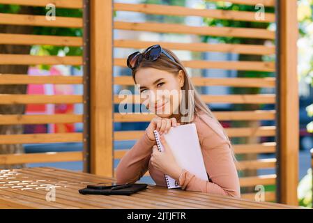 Die junge hübsche Frau hält ein Notizbuch in den Händen und sitzt an einem Tisch in einem Straßencafé oder einem Cavoring Stockfoto