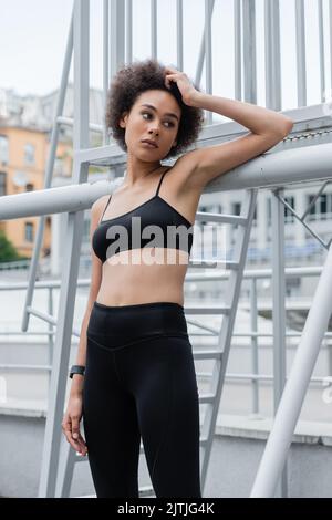 Nachdenkliche afroamerikanische Sportlerin, die lockiges Haar berührt und auf das Stadion blickt Stockfoto