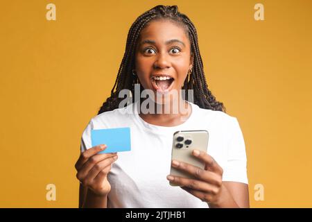 Bild von aufgeregt überrascht afroamerikanischen hübsche Frau posiert isoliert über gelbe Wand Hintergrund mit Handy halten Kreditkarte Stockfoto