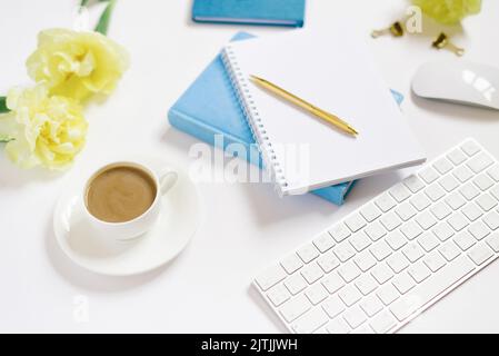 Büroarbeitsplatz mit weißem Schreibtisch. Draufsicht von oben mit Tastatur, Notizblock, Blumen und Bleistift mit einer weißen Tasse Kaffee auf einem Teller. Flacher Surfa Stockfoto