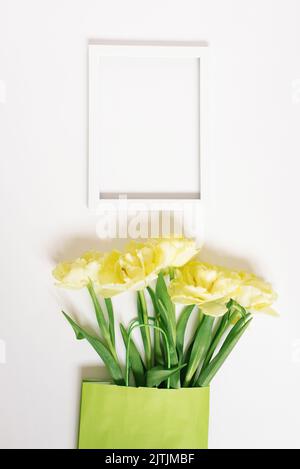 Grüner Papierbeutel mit Frühlingsblumen aus gelben Tulpen und einem weißen leeren Textrahmen auf weißem Hintergrund. Flaches Layout, Draufsicht, Kopierbereich, Modellaufbau Stockfoto
