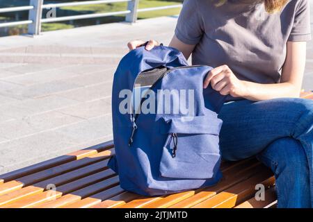 Frauenhänden ziehen einen orangen Schulrucksack mit Schulbedarf, Büchern, einem Büro, einem Laptop auf eine Parkbank. Das Konzept von Back to School. Stockfoto