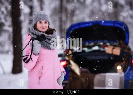 Eine Frau in einem winterverschneiten Wald im Kofferraum eines Autos, das mit Weihnachtsdekor dekoriert ist. Eine Fotografin hält eine Kamera in den Händen. Stockfoto