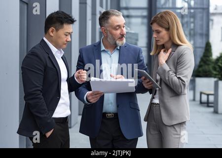 Diverse Business-Gruppe von drei Kollegen Arbeitnehmer, aufgebracht und enttäuscht, Ergebnis des Finanzberichts und Vertragsdokument, Geschäftsleute außerhalb Bürogebäude in Geschäftsanzüge reden. Stockfoto