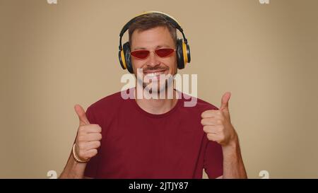 Fröhlicher, gutaussehender, entspannter Mann mit purpurfarbener Frisur, der über Kopfhörer Musik hört und Disco tanzt, die sich lustig mit ausdrucksstarken gestikulierenden Händen herumtollt. Erwachsene stilvolle männlichen Kerl drinnen Stockfoto