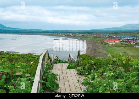 Blick auf die Bucht von Juschno-Kurilsk auf der Insel Kunaschir von einem hohen Kap, im Vordergrund ein hölzerner Bürgersteig mit einer Leiter Stockfoto