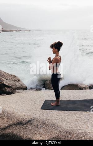 Die ganze Länge einer schlanken Frau, die gegen die Wellen meditiert. Seitenansicht einer jungen Frau, die im Freien am Meer Yoga praktiziert. Stockfoto