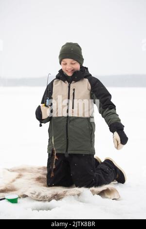 Fröhlicher Junge in warmer Kleidung, der beim Angeln auf dem gefrorenen See kniet Stockfoto