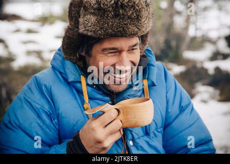 Glücklicher reifer Mann in warmer Kleidung, der Tee in Holzbecher trinkt Stockfoto