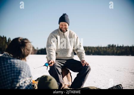 Glücklicher reifer Mann hält Angelrute sitzen mit Freunden während sonnigen Tag im Winter Stockfoto