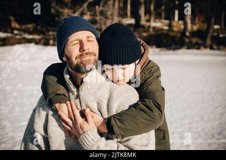 Junge in warmer Kleidung umarmt Vater mit geschlossenen Augen an sonnigen Tag im Winter Stockfoto