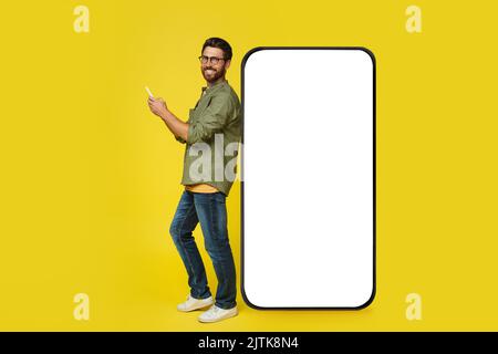 Fröhlicher Mann in lässigem Outfit, der sich auf einem riesigen Smartphone mit weißem, leeren Bildschirm stützt, Handy benutzt und lächelt, Mockup Stockfoto