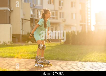 Kleines Mädchen auf Rollschuhe in einem Park. Ein Kind reitet auf Rollschullauffahrten mit ausgestreckten Armen vor dem Hintergrund der grünen Pflanzen im Park Stockfoto