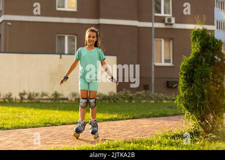 Kleines Mädchen auf Rollschuhe in einem Park. Ein Kind reitet auf Rollschullauffahrten mit ausgestreckten Armen vor dem Hintergrund der grünen Pflanzen im Park Stockfoto